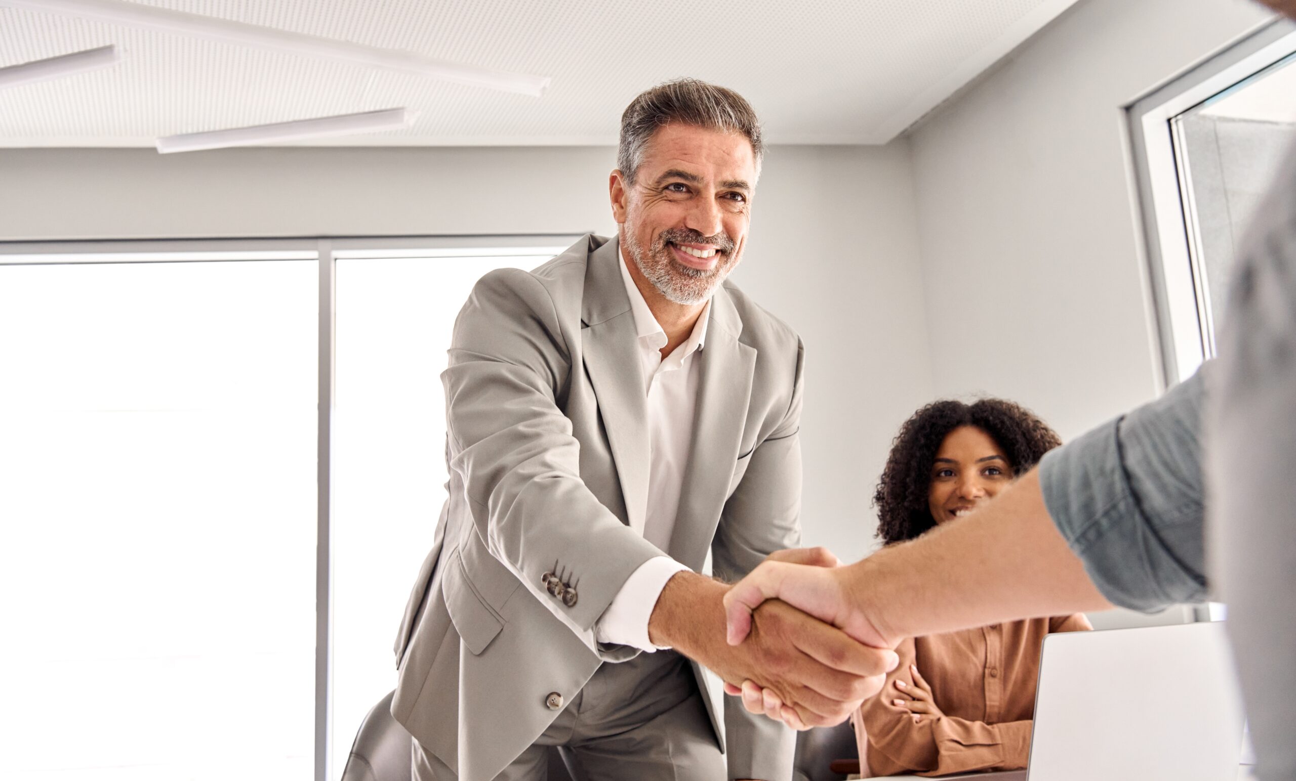 A smiling recruiter in a business suit shaking hands with a candidate during a job interview, symbolizing confidence, suitability, and selection.