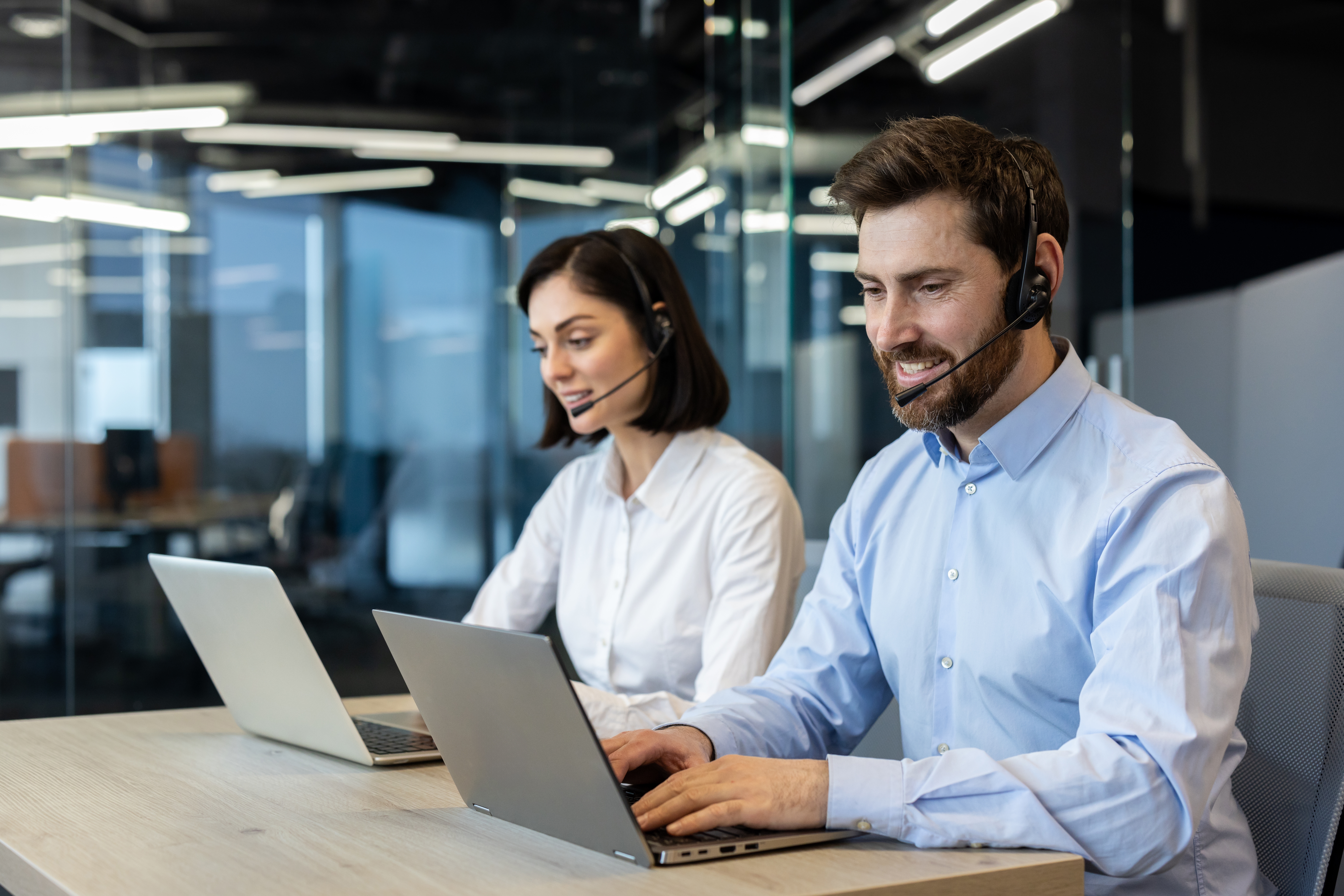 Two customer support representatives wearing headsets and working on laptops in a modern office environment.